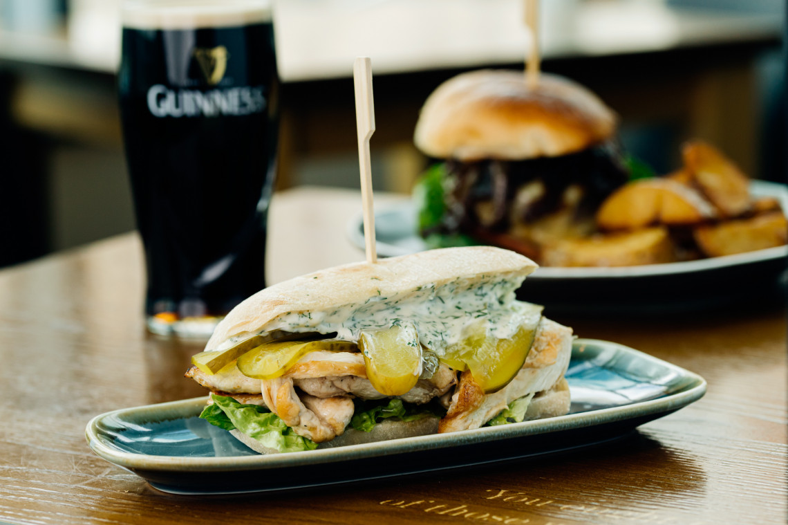 A plate with a chicken ciabatta, gherkins and mayonnaise. Behind it, a pint of Guinness and a burger and wedges blurred behind that.